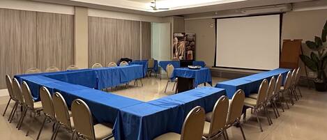 Un salón de conferencias con una larga mesa, sillas, pantalla de proyector y ventiladores de techo.