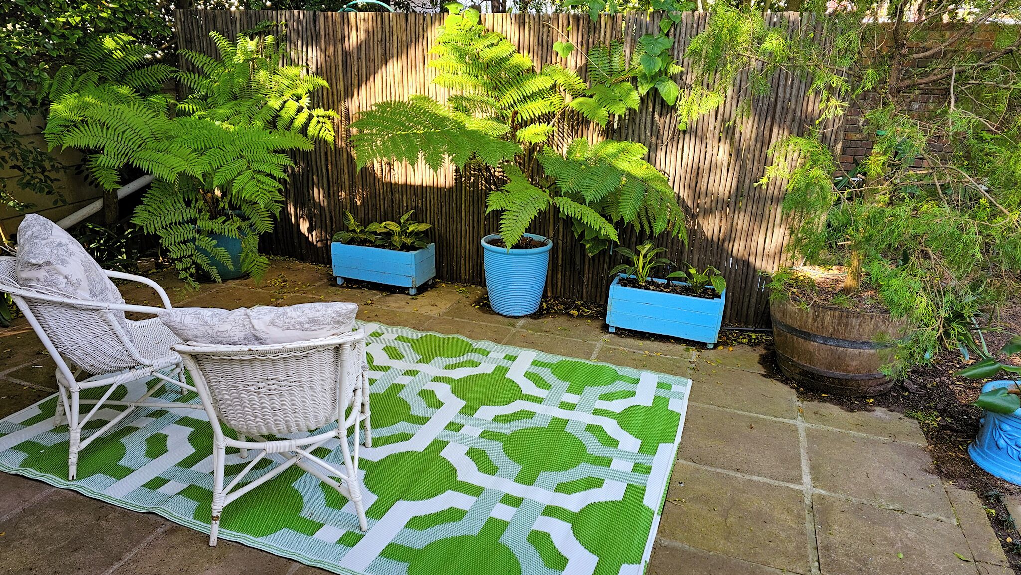 A patio area with a wicker chair, a patterned rug, and potted plants against a wooden fence.