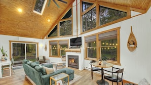 A spacious living area with a fireplace, large windows, and a high ceiling.