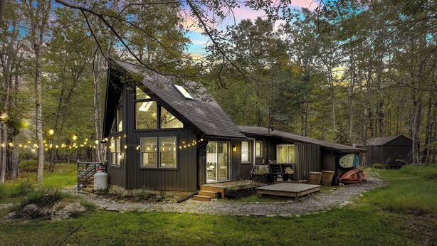 A modern house with large windows, a deck, and a small shed in a forested area.