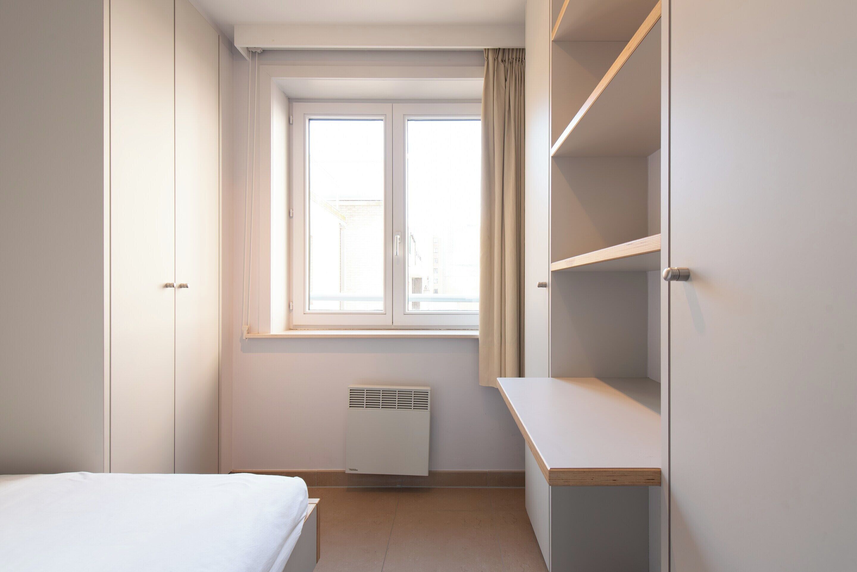 A bedroom with a bed, a window with curtains, a radiator, and a desk.
