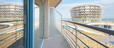 A balcony with a view of two distinctive buildings with a geometric design, a clear sky, and a body of water in the background.