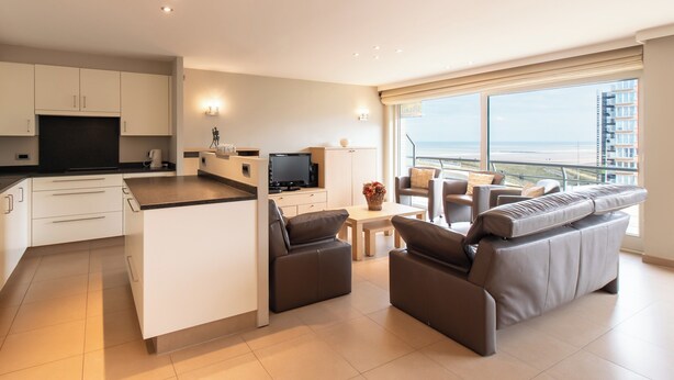 A modern living area with a kitchen, a sofa set, a dining table, and a view of the sea.