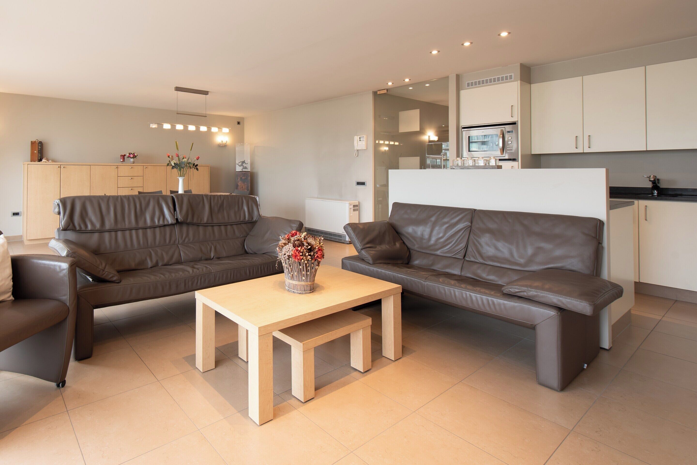 A modern living room with two brown leather sofas, a wooden coffee table, and a kitchen area with white cabinets and a microwave.