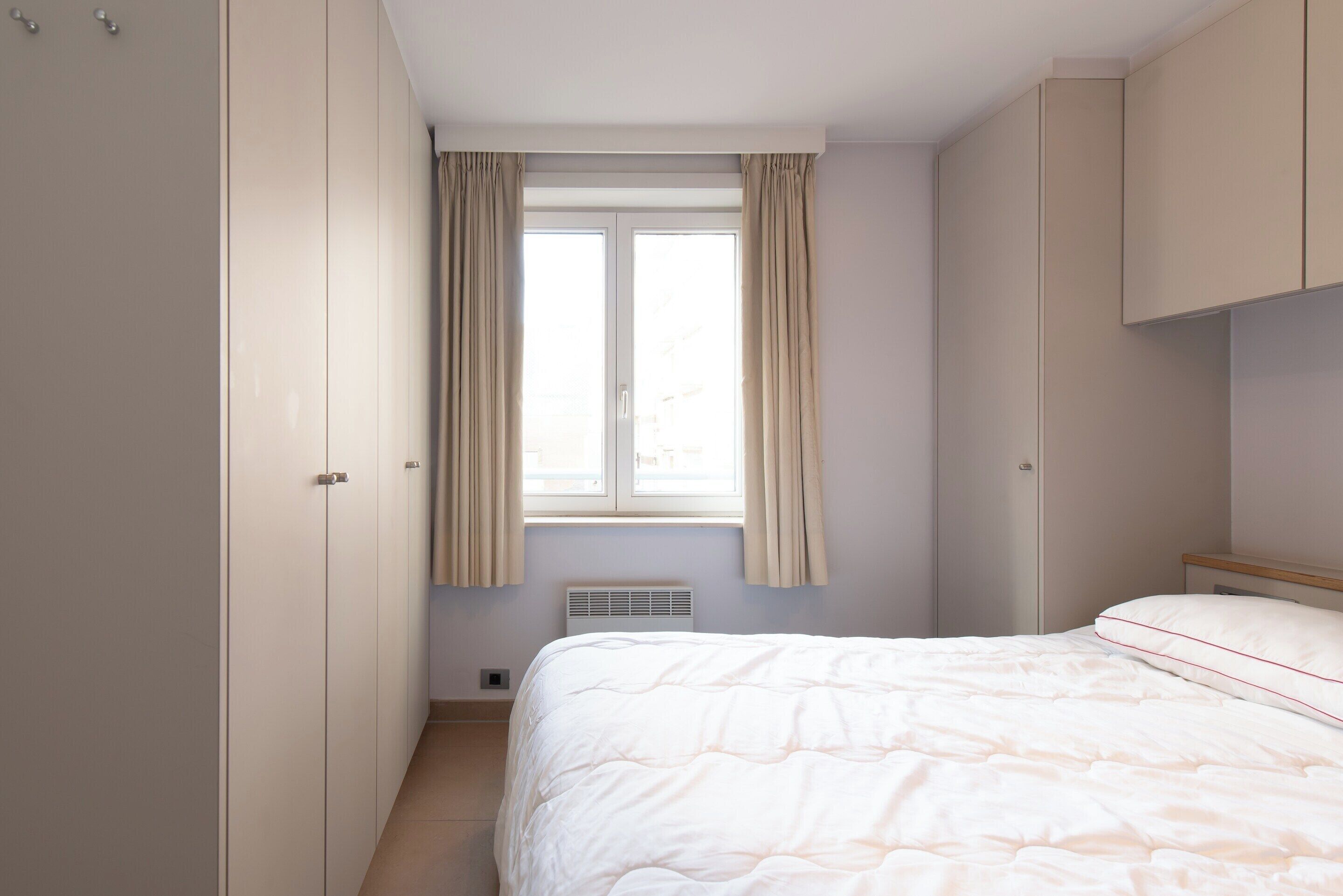 A bedroom with a bed, a window with curtains, a radiator, and a wardrobe.