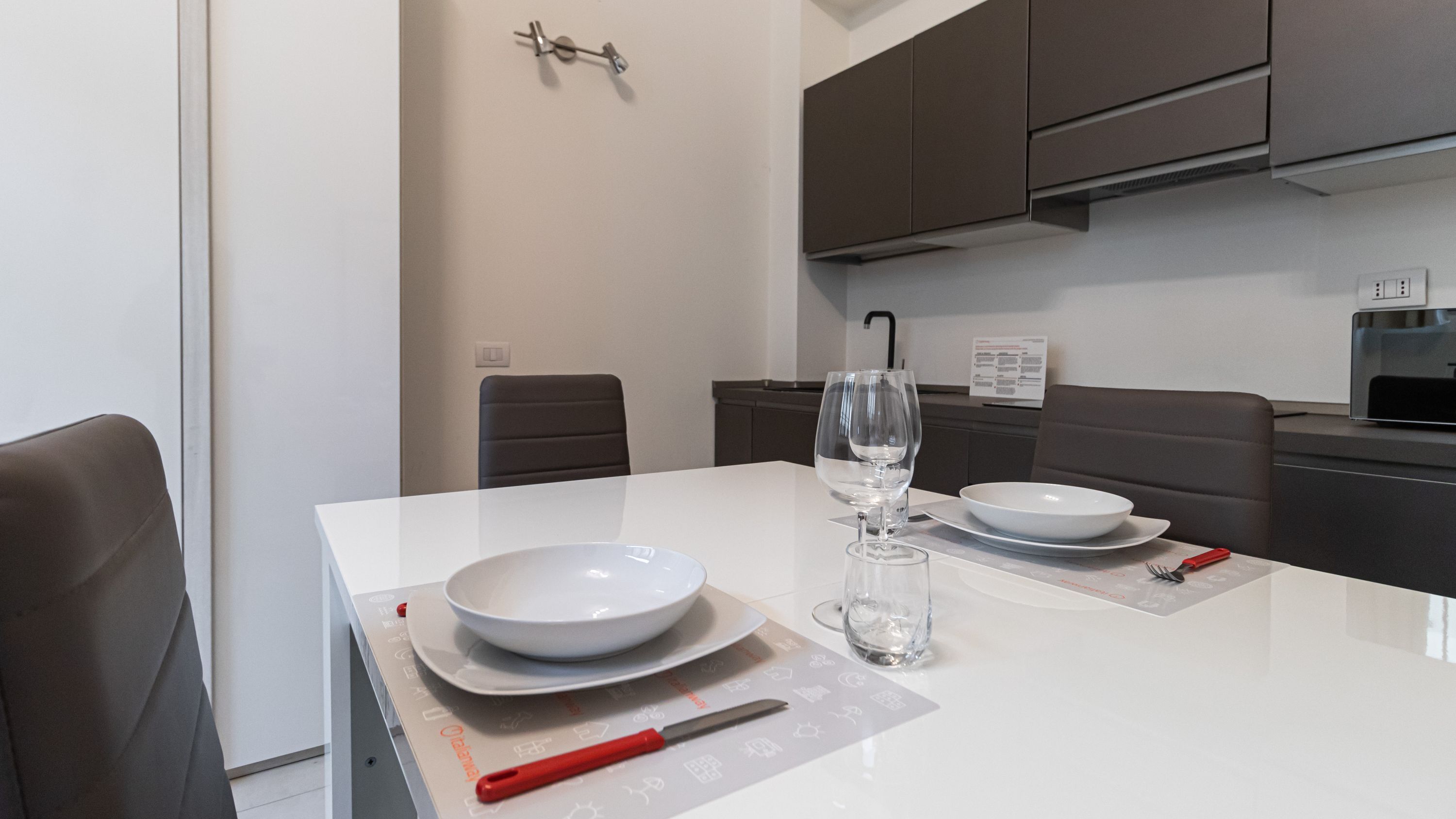 A modern kitchen with white countertops, dark cabinets, and a dining table set with plates, a wine glass, and cutlery.
