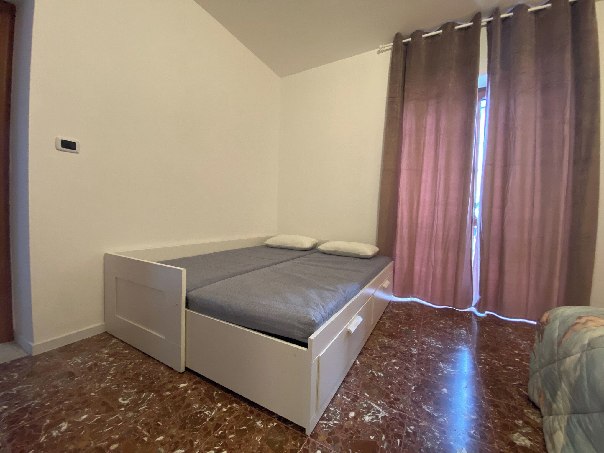 A small, single-bed room with a simple bed, a small bedside table, and a window with curtains.