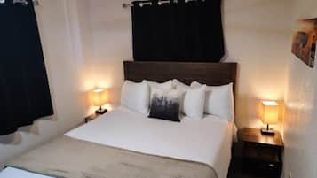 Un lit bien fait, avec du linge de lit blanc et une tête de lit sombre, flanqué de deux tables de chevet avec des lampes, dans une pièce dotée d’un ventilateur de plafond et de rideaux noirs.
