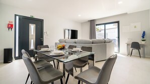 A modern dining area with a glass table, grey chairs, and a sofa in the background.