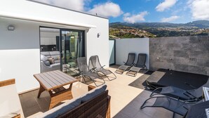 A rooftop terrace with a glass door leading to a kitchen, lounge chairs, a table, and a view of a hilly landscape.