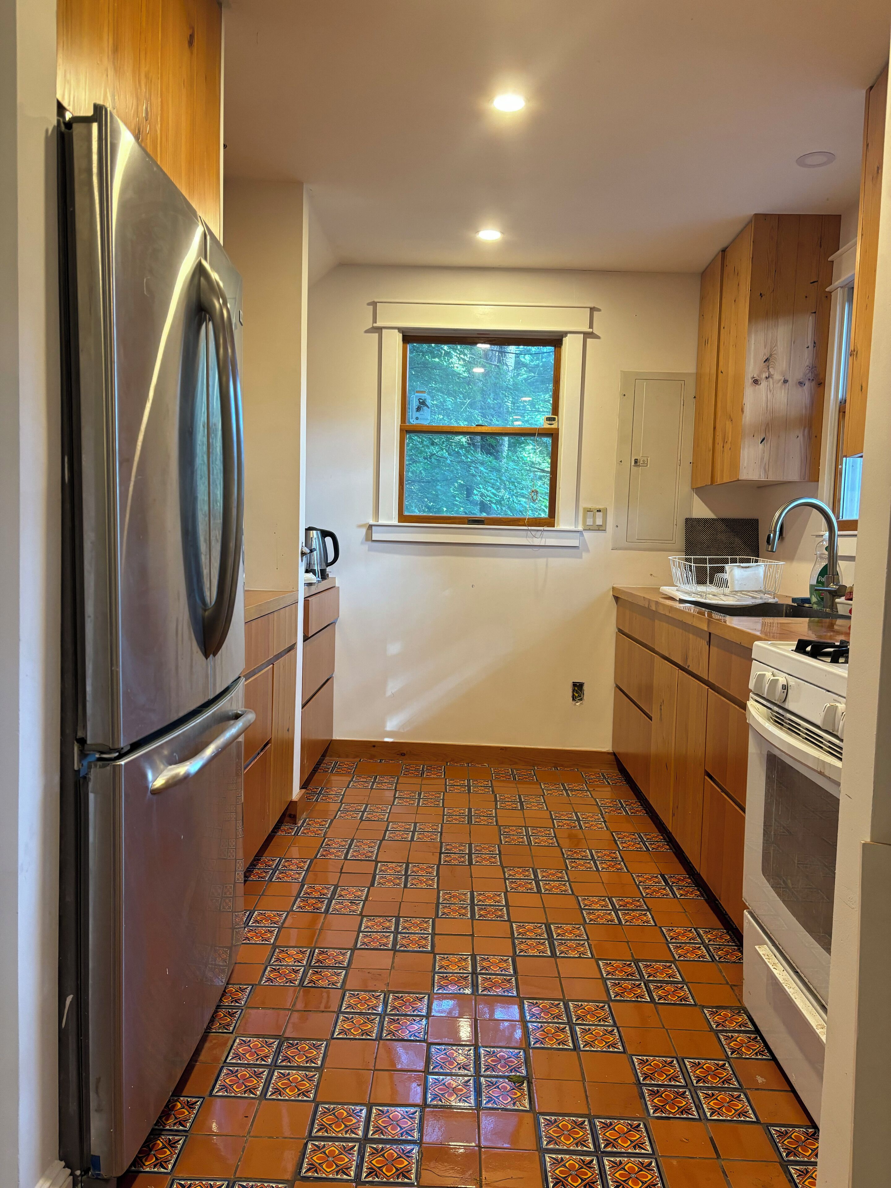 Una cocina compacta con refrigerador de acero inoxidable, ventana con vistas a árboles, piso de baldosas y armarios de madera.