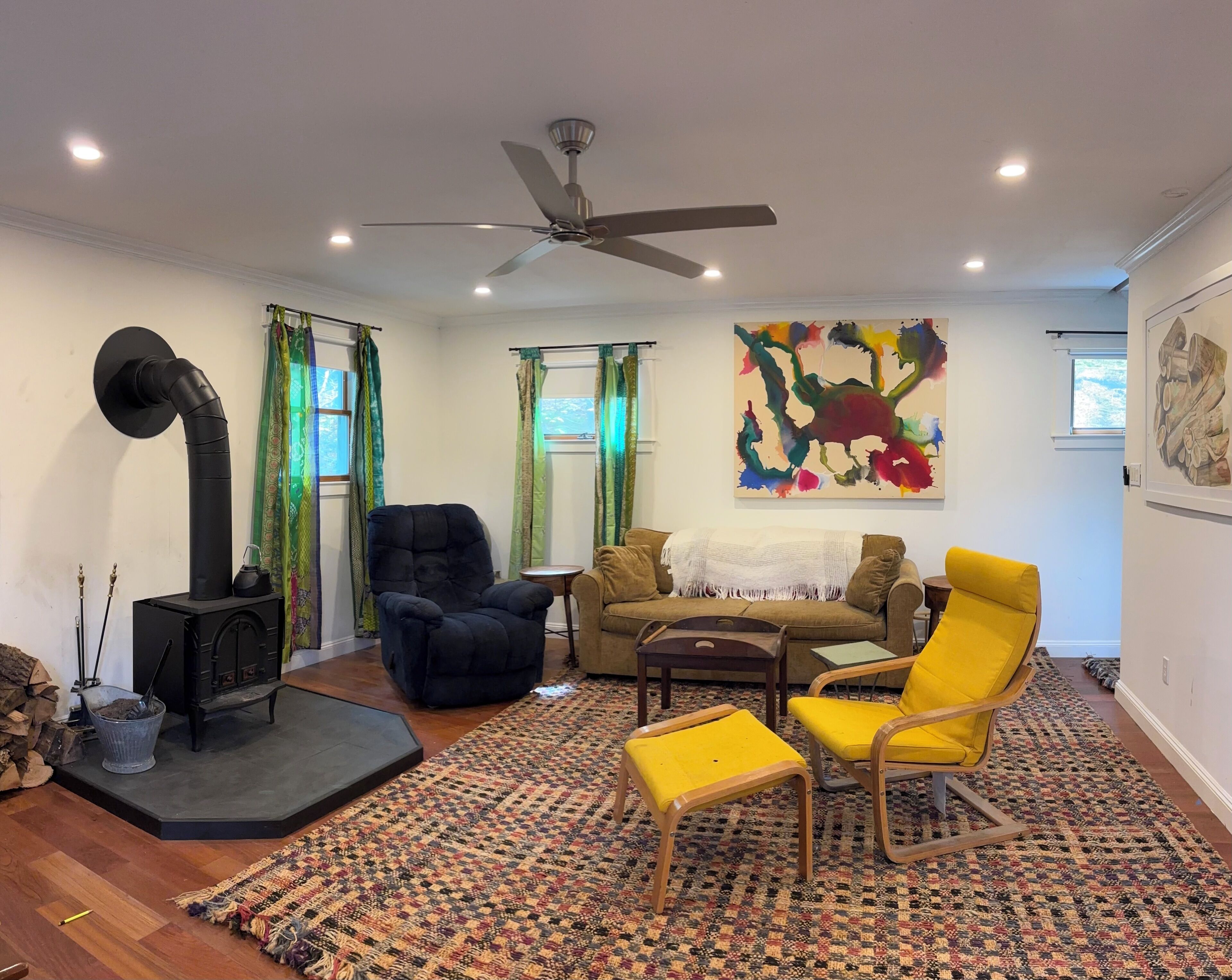 Una sala de estar con chimenea, ventilador de techo, sofá, sillón amarillo y alfombra estampada.