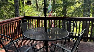 Una mesa redonda de metal con cuatro sillas, una planta colgante y una bandera en una terraza de madera con barandilla.