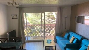 A living room with a blue sofa, a small table, a TV, and a sliding glass door leading to an outdoor area with a fence and chairs. - Lovely apartment near city center (Canet-en-Roussillon)