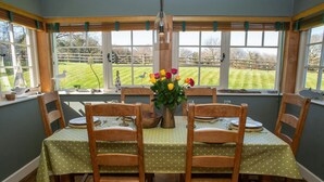 A dining room with a table set for four, surrounded by wooden chairs, a vase of flowers, and a view of a grassy yard through large windows. - Ol y Don, Newport - Three Bedroom Cottage, Sleeps 6 (Newport)