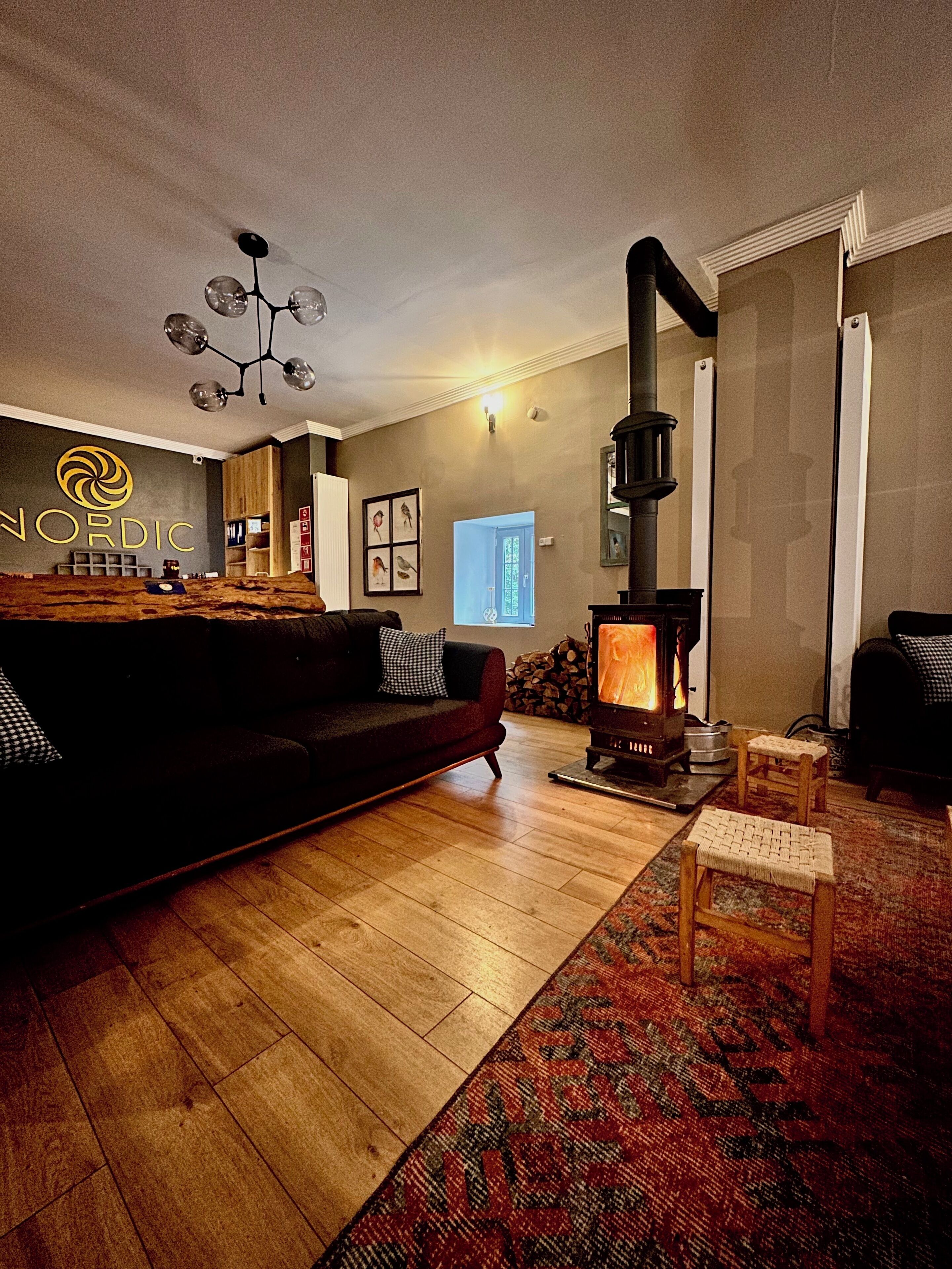 A cozy living room with a wooden floor, a dark-colored sofa, a small wooden table, and a wood-burning stove. There is a sign with the word "NORDIC" on the wall.