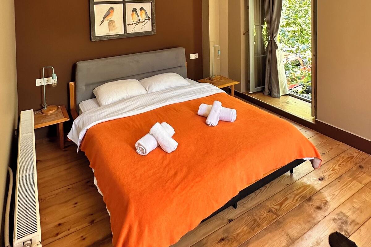 A bedroom with a wooden floor, a bed with an orange bedspread, white pillows, a framed picture of birds on the wall, and a window with curtains.
