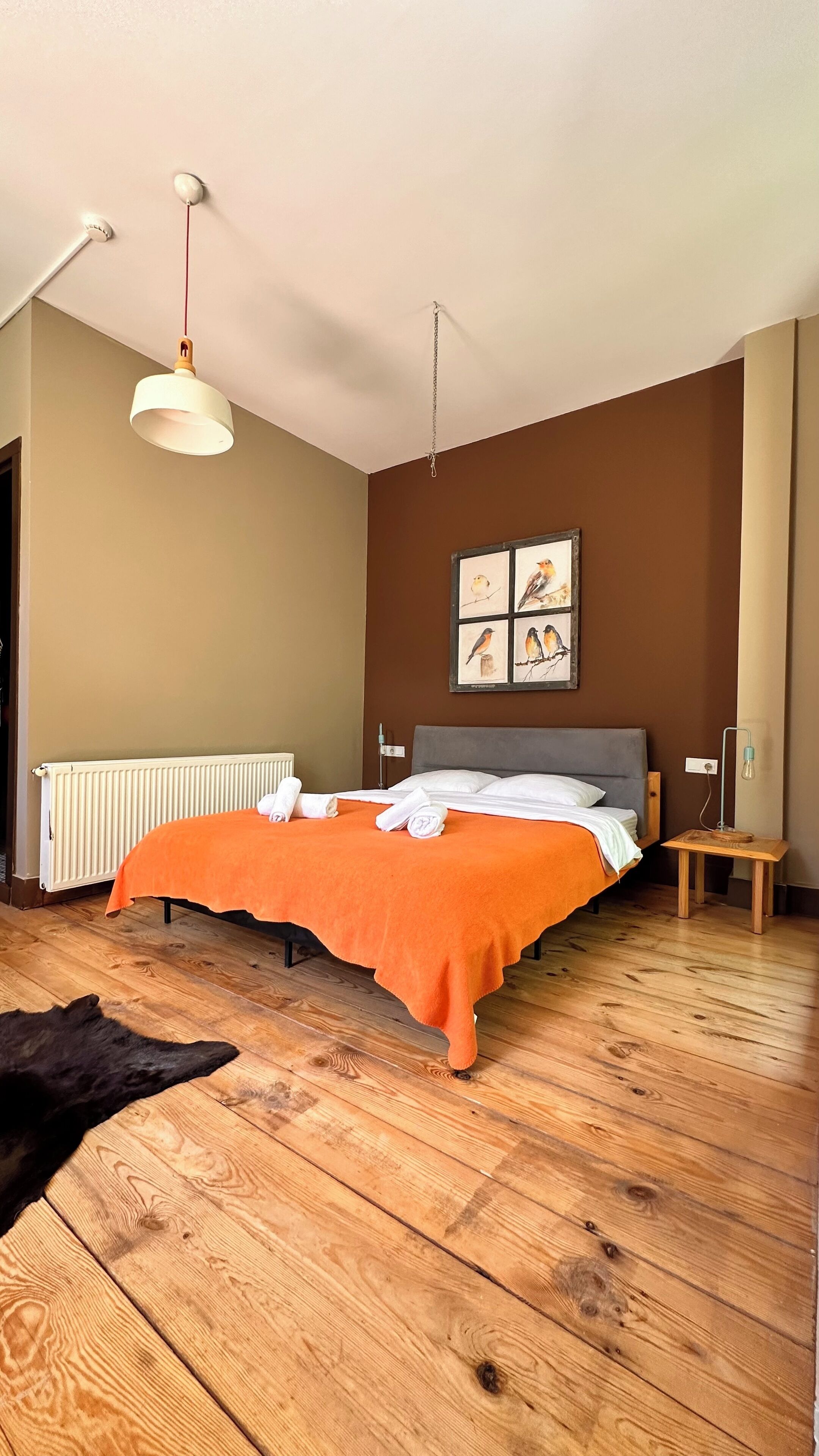 A bedroom with a bed, a radiator, a wooden floor, and a hanging light fixture.