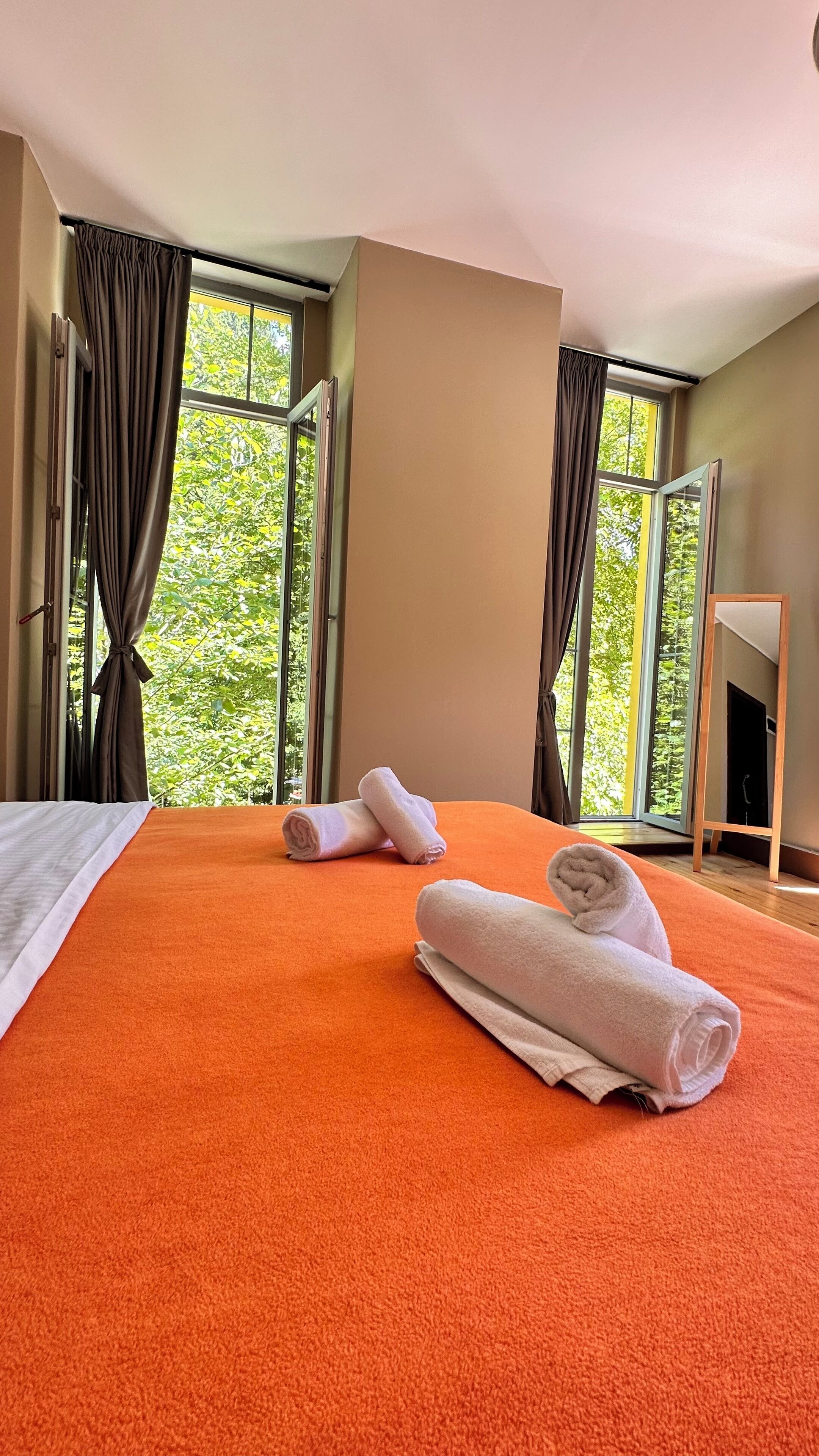 Ein Hotelzimmer mit einem großen Bett, zwei Fenstern mit Vorhängen und einem Holznachttisch.