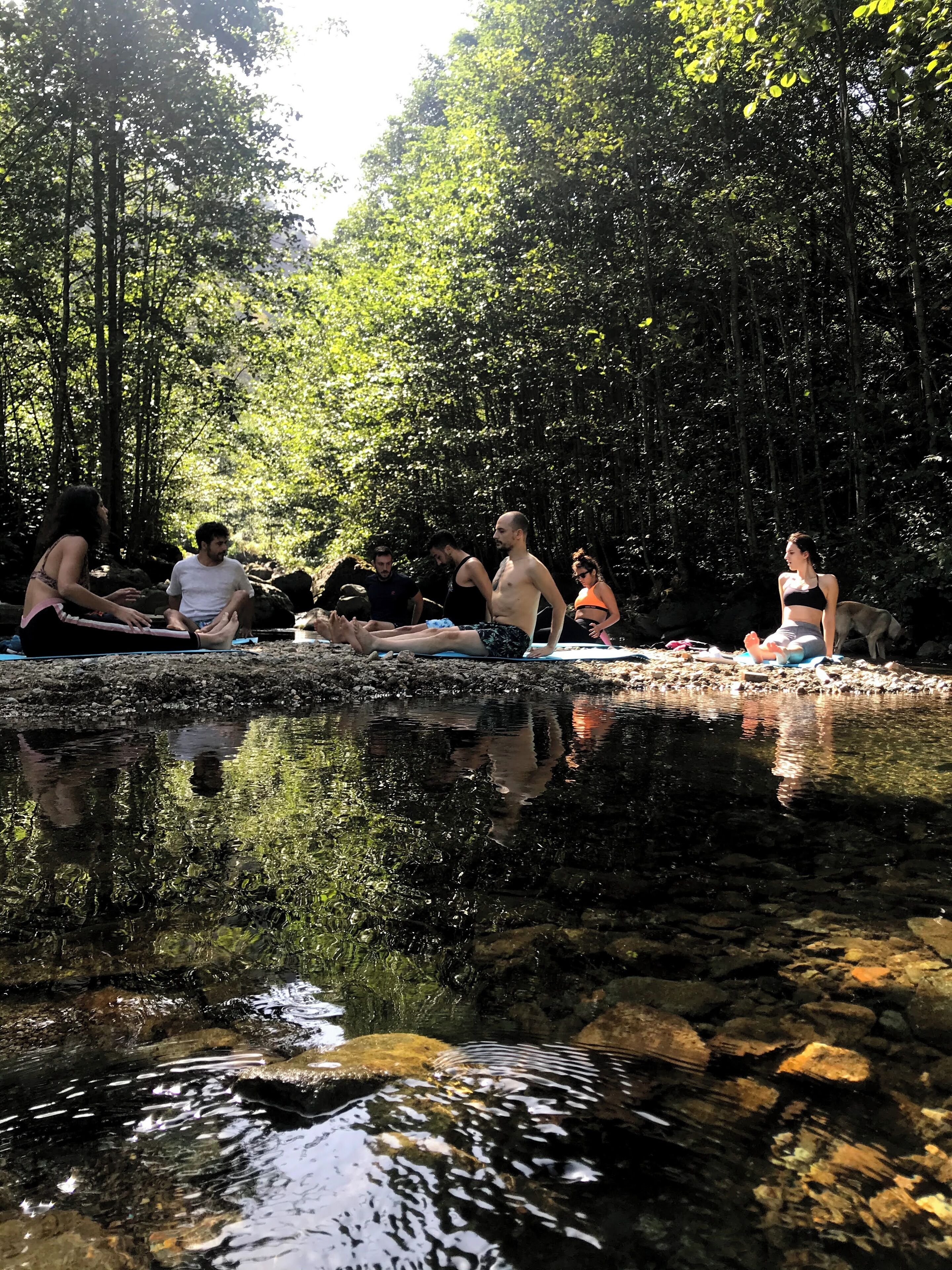 A group of people sitting by a clear stream in a forested area.