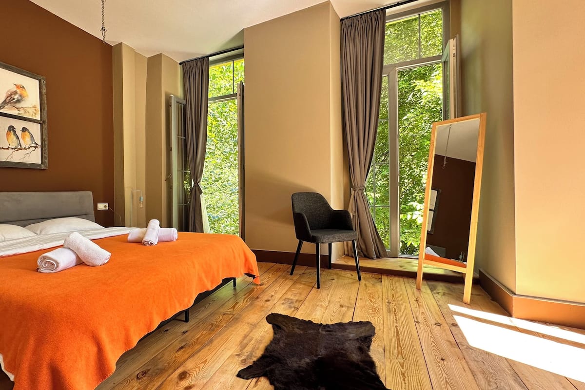 A bedroom with a bed, a chair, a large window, and a mirror.