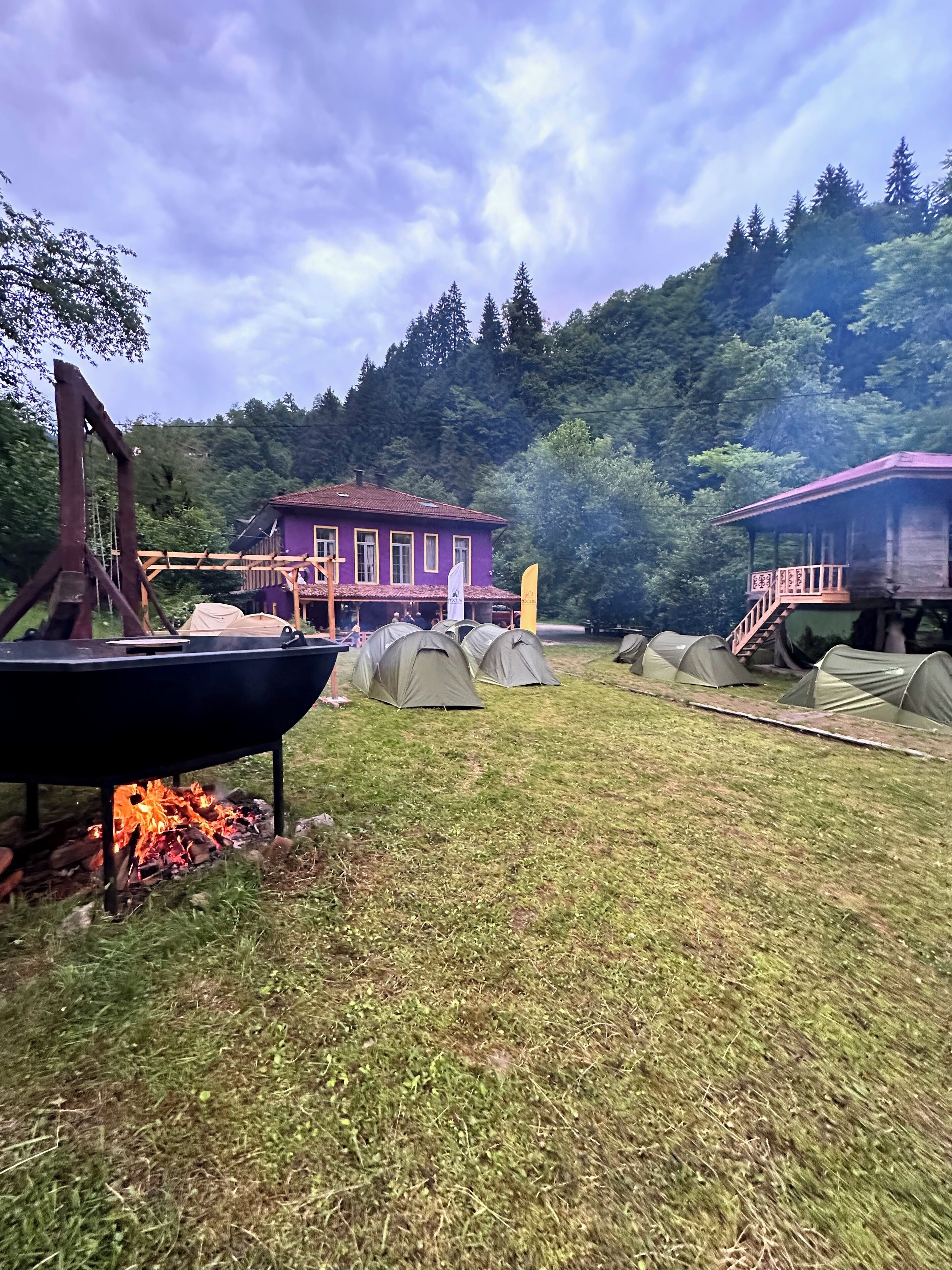A rustic outdoor area with a purple building, a fire pit, and several tents.
