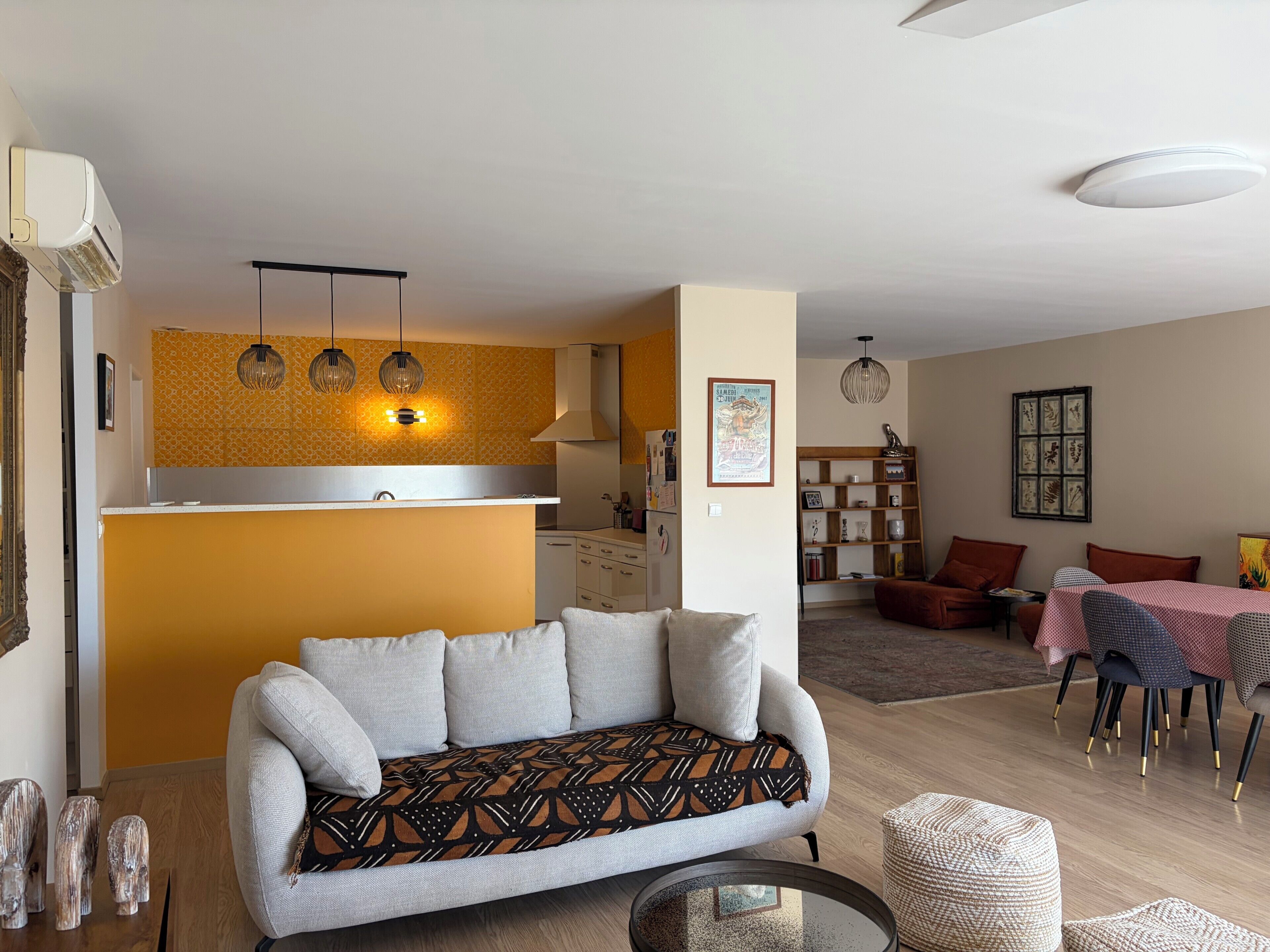 Un salon avec un canapé, un coin repas avec une table et des chaises, un coin cuisine avec un comptoir et des luminaires suspendus, ainsi qu’un luminaire fixé au plafond.