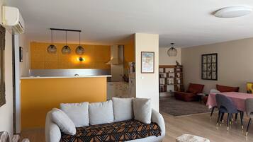 Un salon avec un canapé, un coin repas avec une table et des chaises, un coin cuisine avec un comptoir et des luminaires suspendus, ainsi qu’un luminaire fixé au plafond.