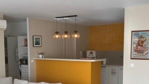A modern living room with a sofa, a wooden dining table, and a kitchen area with a yellow accent wall and pendant lights. - Stylish Apartment - 5 Min Walk to Center (L'Isle-sur-la-Sorgue)