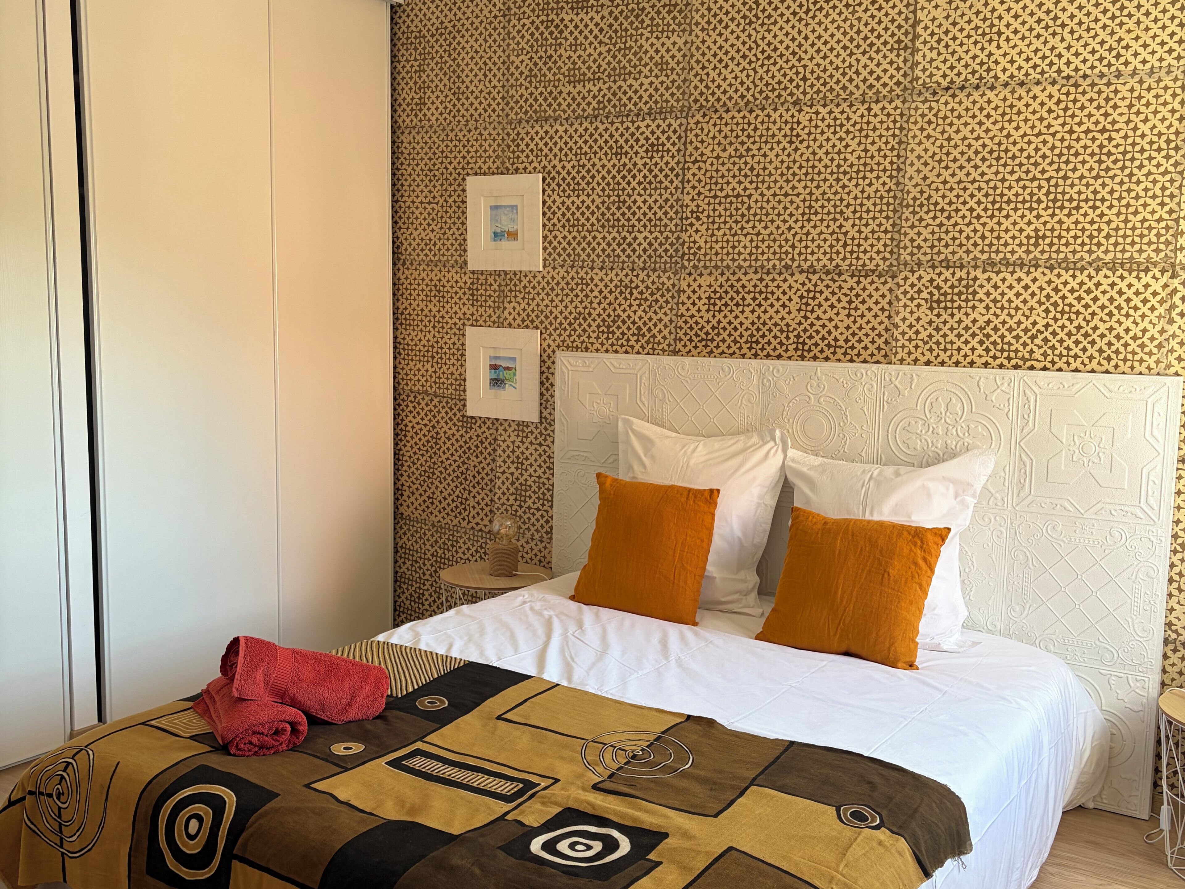 Une chambre d’hôtel avec un mur orné d’un motif, un lit avec des oreillers blancs et orange, une couverture de lit marron à motifs et une serviette rouge posée sur le lit.