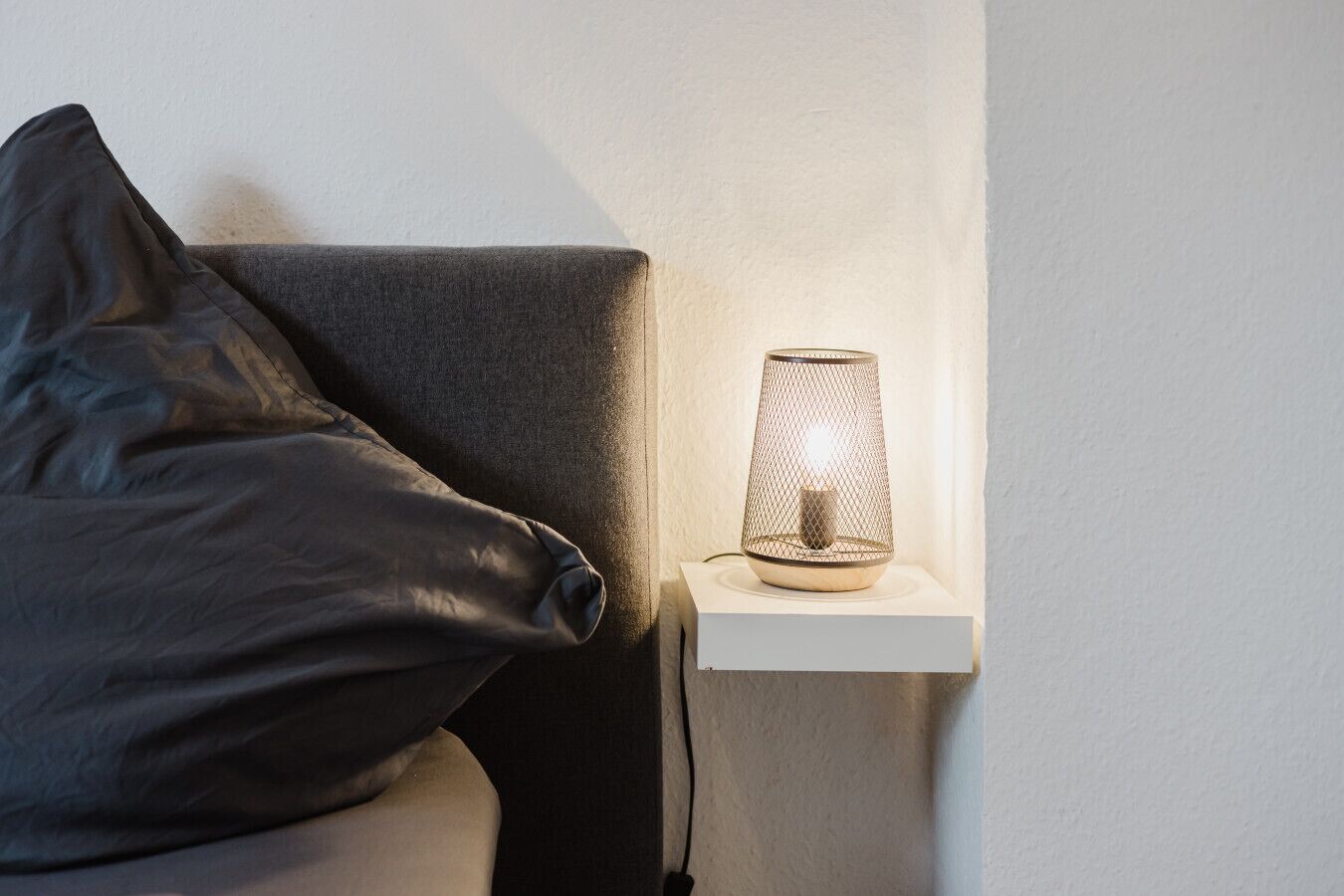 Un comodino con una lampada, un cuscino grigio scuro e una parete bianca.