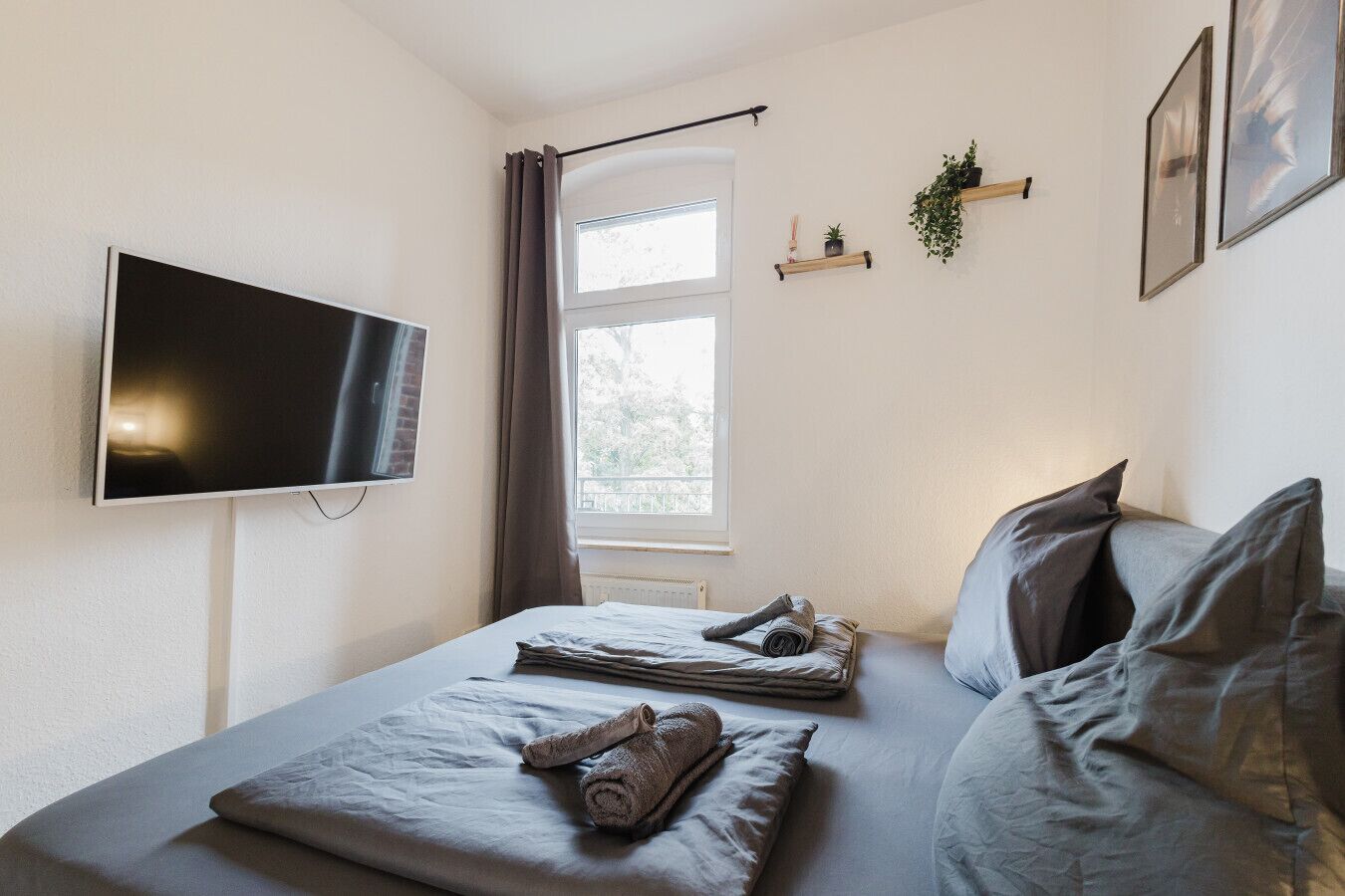 Un dormitorio con cama, televisor, ventana y una planta en un estante.