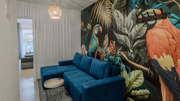 A modern living room with a blue sectional sofa, a round coffee table, and a vibrant tropical mural. - Passau Apartment|Singles-Couples-Groups|Central (Passau)