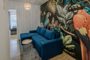 Un salon moderne avec un canapé d’angle bleu, une table basse ronde et une fresque murale aux couleurs vives évoquant les tropiques.