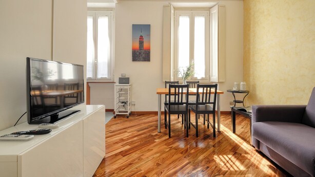A living room with a flat-screen TV, wooden flooring, a dining table with chairs, a sofa, and a painting of a tower on the wall. - JOIVY Bright flat near the city centre  (Milan)