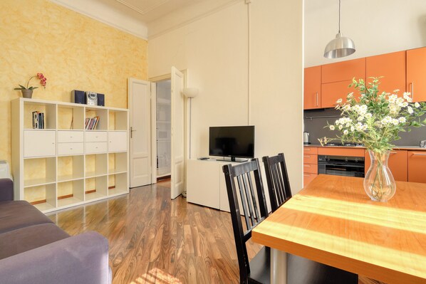 A modern kitchen with orange cabinets, a dining table with chairs, a TV, and a bookshelf.
