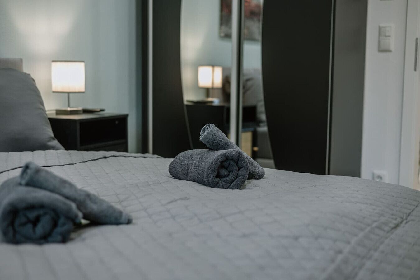 Ein ordentlich bezogenes Bett mit einer grauen Tagesdecke und zwei aufgerollten Handtüchern.