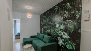 Un salon moderne avec un canapé d’angle vert, une table basse ronde et une fresque murale représentant un feuillage tropical.