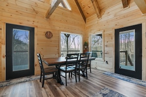 A wooden cabin interior with a dining table and chairs, large windows, and a clock on the wall.