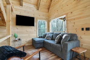 Ein gemütliches Holzhaus-Interieur mit einer Eckcouch, einem flachbildfernseher an der Wand und großen Fenstern mit Blick ins Freie.