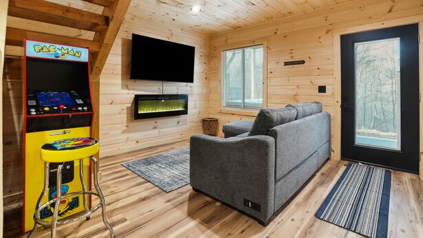 A cozy cabin interior with a gray sofa, a Pac-Man arcade machine, a flat-screen TV, and a large window with a view of trees. - Timberline Retreat | Hocking Hills, Hot Tub (Creola)