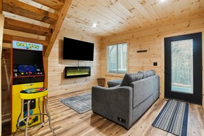 A cozy cabin interior with a gray sofa, a Pac-Man arcade machine, a flat-screen TV, and a large window with a view of trees. - Timberline Retreat | Hocking Hills, Hot Tub (Creola)