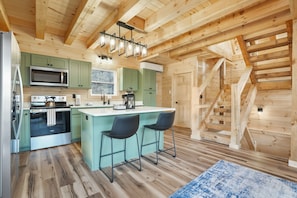 Une cuisine moderne avec des poutres en bois, des armoires vertes, un îlot central doté de tabourets de bar et un escalier menant à l’étage supérieur.