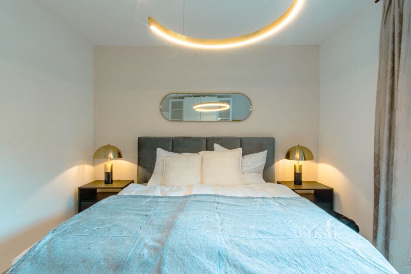 Ein ordentlich bezogenes Bett mit einer hellblauen Tagesdecke, zwei Nachttischlampen und einem Spiegel über dem Bett.
