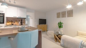 Una cocina moderna con armarios blancos y encimera de madera, una zona de comedor con una silla azul, un televisor sobre un mueble de madera y una planta en una mesita auxiliar.