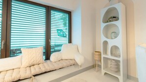Una sala de estar moderna con un sofá blanco, una estantería alta de color blanco y elementos decorativos, y ventanas grandes con persianas.