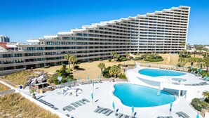 Un hôtel doté de plusieurs piscines, un grand bâtiment en arrière-plan et un espace aménagé avec des chaises longues et des parasols.