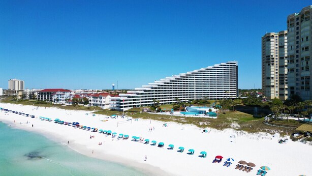 Un complexe hôtelier de bord de mer doté d’une longue plage de sable, de nombreux parasols et d’un grand bâtiment principal comprenant plusieurs étages.