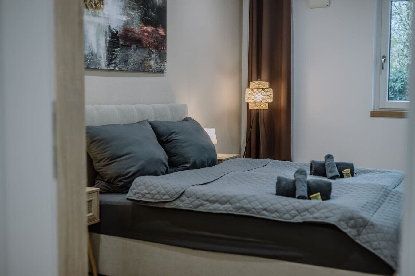 Un lit bien fait, recouvert d’une couverture grise matelassée, avec deux oreillers et une lampe de chevet.
