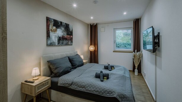 Un dormitorio moderno con una cama grande, una mesita de noche con lámpara, un cuadro en la pared, una ventana con cortinas y un televisor montado.